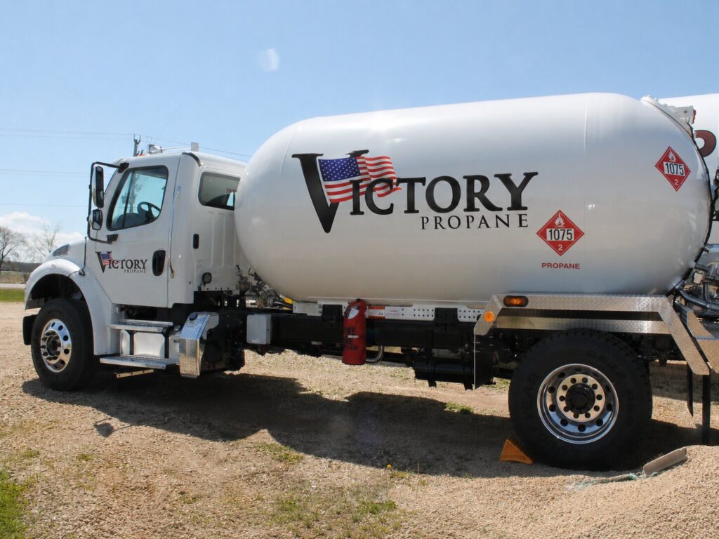 Victory Propane delivery truck parked on gravel, driver-side profile showing the white tank with the American flag Victory Propane logo and 1075 propane placard.
